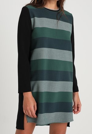 Femme portant une robe à manches longues avec de larges rayures horizontales noires, vert foncé, bleu marine et grises, dotée d'évents latéraux à l'ourlet.