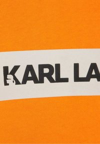 Camiseta de algodón naranja con una banda blanca en el centro donde destacan grandes letras negras que dicen "KARL LAGERFELD".