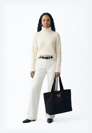 Sieviete krēmkrāsas augstu uzrullēta kakla džemperī un baltās biksēs, turot lielu melnu roku somu ar sarkanu sirsniņas logotipu, stāvot pret baltu fonu.