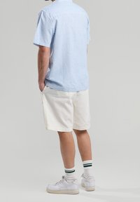 Modrá a bílá pruhovaná košile s krátkým rukávem a límečkem, v kombinaci s béžovými kraťasy a bílými teniskami se zelenými detaily, pohled zezadu.