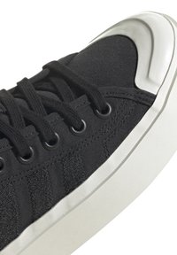 Baskets noires en toile avec une tige texturée, lacets noirs et œillets ronds. La coque de protection est blanche brillante et la semelle est plate et blanche.