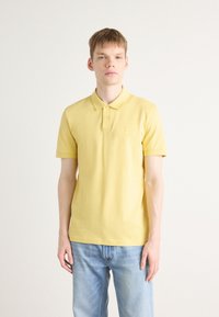 Gul polo-skjorte laget av bomull, med kragehals, korte ermer, og en liten logo-patch på brystet. Brukt sammen med blå jeans.