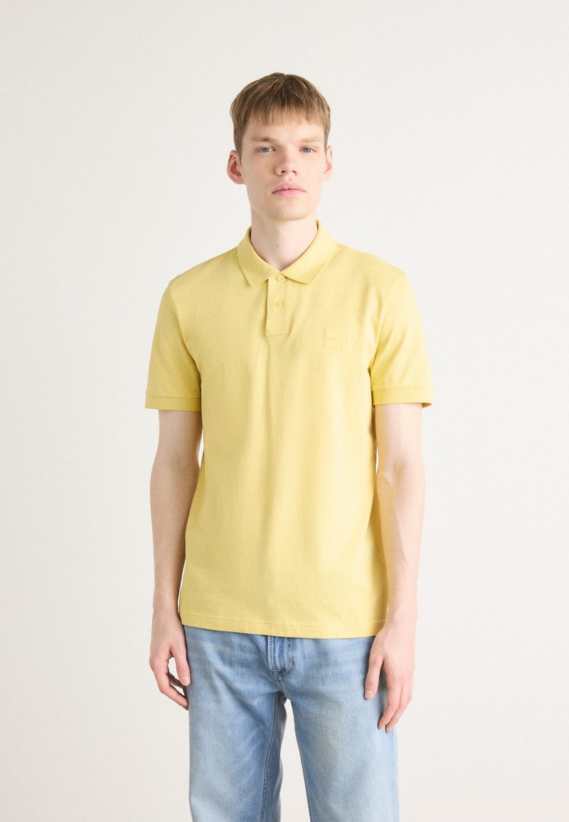 Gul polo-skjorte laget av bomull, med kragehals, korte ermer, og en liten logo-patch på brystet. Brukt sammen med blå jeans.