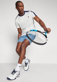 Hvit og marineblå stripete treningsskjorte, blå shorts, hvite tennissko med marineblå detaljer, holder en tennisracket, stående på ett ben.