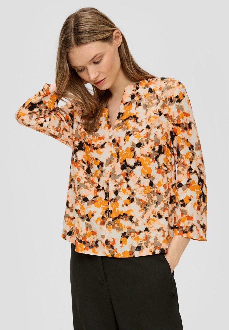 Vrouw met lichtbruin haar draagt een losse oranje, zwarte en beige gebloemde blouse met zwarte broek, hand in de zak, en kijkt naar beneden.