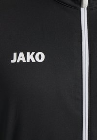 Schwarze Jacke mit einem weißen Reißverschluss und dem "JAKO"-Logo in weißer Schrift auf der linken Brustseite. Glattes Material mit taillierter Passform.
