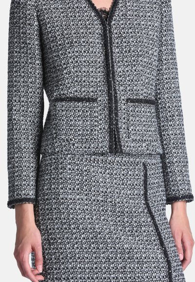 Femme portant une veste en tweed noir et blanc et une jupe assortie avec des bordures noires et deux poches avant sur la veste.