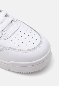 White leather sneaker with perforated toe box, flat laces, contrasting stitching, rounded shape, and rubber sole. Smooth texture.