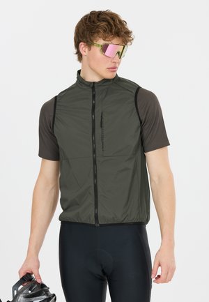 Junger Mann mit reflektierenden Sonnenbrillen, einer olivgrünen Weste über einem schwarzen Radtrikot und schwarzen Radhosen, der einen Helm hält.