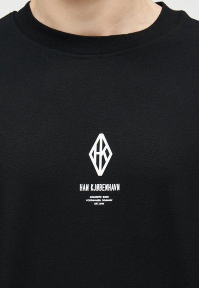 Μαύρο μπλουζάκι με λευκό λογότυπο Han Kjøbenhavn και κείμενο «Concrete Born Copenhagen Denmark Est. 2008» τοποθετημένο στο κέντρο κάτω από το λαιμόκοψη.