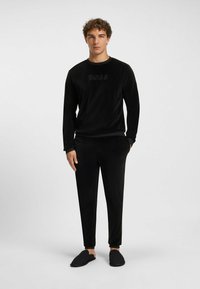 Tuta in velluto nero caratterizzata da una maglietta a maniche lunghe con logo "BOSS", pantaloni abbinati e scarpe nere senza lacci. Texture liscia e design aderente.