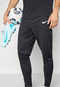 Sorte atletiske bukser med en tætsiddende pasform og elastiske manchetter. En lyseblå fodboldstøvle med en fremtrædende swoosh holdes ved siden af en mønstret fodbold.