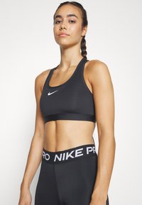 Black sports bra made of stretchy fabric with a round neckline, racerback design, and white Nike logo. Worn with black Nike Pro waistband.