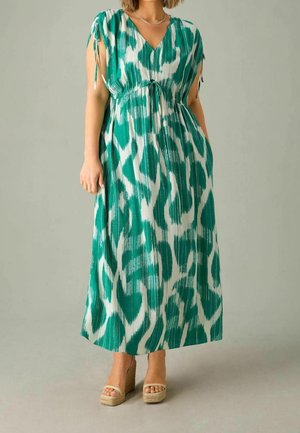 Frau trägt ein ärmelloses Maxikleid mit grün-weißem Muster, das an den Schultern Kordelzüge hat, sowie beige Keilsandalen.