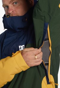 Chaqueta multicolor con paneles en azul marino, verde y amarillo; incluye bolsillos con cremallera y un cuello alto con características ajustables.