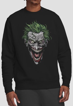 Mann trägt schwarzen Pullover mit detailliertem, unheimlichem Gesicht einer Figur mit grünen stacheligen Haaren, blasser Haut, weit geöffneten Augen und einem großen, offenen Mundgrinsen.