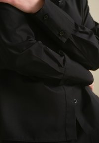 Zwarte button-up blouse met lange mouwen, gemaakt van gladde stof. Heeft een standaard kraag en zwarte knopen op de mouwen en aan de voorkant.