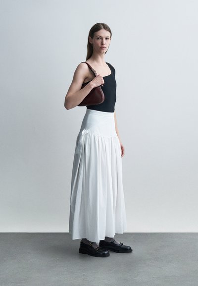 Γυναίκα με μαύρο αμάνικο τοπ, μακριά λευκή φούστα, μαύρα παπούτσια, κρατώντας σκούρο καφέ τσάντα ώμου πάνω σε απλό ανοιχτό φόντο.