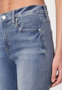 Primer plano de la sección media vistiendo jeans de mezclilla azul claro con bolsillos frontales y de reloj visibles, y una mano apoyada al lado.