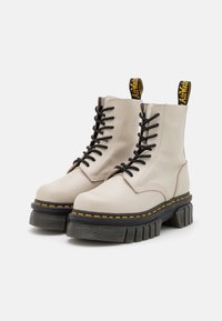 Botas de tobillo beige con cordones, suela de goma, cordones negros, costuras amarillas y una pestaña para tirar. Diseño de plataforma gruesa con un acabado texturizado.