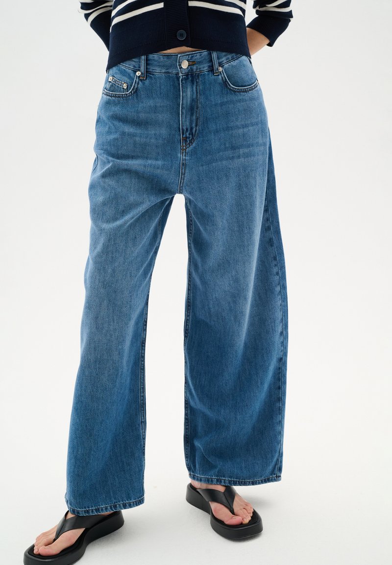 Jeans anchos de color azul, hechos de denin, con cintura alta, bolsillos delanteros, cierre de botón y detalles de dobladillo sin acabar. Combinados con chanclas negras.