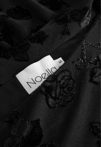 Fekete anyag, kiemelkedő virágos hímzéssel, sima textúrával. Egy fehér címke látható, amelyen "Noella" és az "S" méret olvasható.