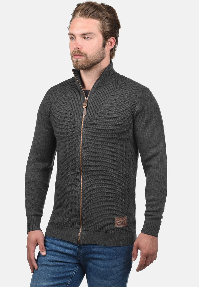Homme barbu portant un pull texturé gris foncé à fermeture éclair intégrale et un jean bleu, debout, les mains détendues le long du corps.