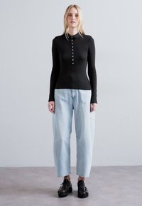 Top lungo a maniche nere a coste con colletto a camicia e sei bottoni, abbinato a jeans chiari larghi e scarpe scure.