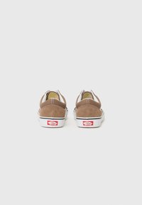 Bruna mockaskateskor med låg skärning, vita gummisulor och en röd Vans-logotyp på hälen. Har elastiska sidodetaljer för en bra passform.
