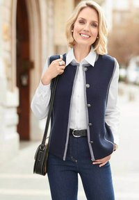 Glimlachende blonde vrouw in een witte blouse, marineblauwe vest met knopen, blauwe jeans, zwarte riem, terwijl ze een zwarte schoudertas buiten draagt.