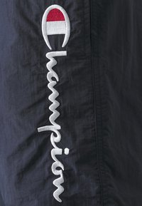 Șorturi sportive din țesătură neagră, cu broderie albă cursivă "Champion" și un emblema roșie și albă în partea de sus. Material texturat.