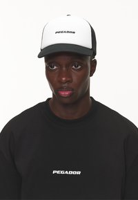 Schwarze Kappe mit weißem Frontpanel und schwarzen Mesh-Seiten, mit "PEGADOR" in fetter Schrift. Schwarzer Sweatshirt mit passendem Logo.