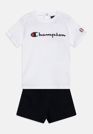 T-shirt bianco in cotone con logo "Champion" nero, abbinato a pantaloni neri. La t-shirt presenta maniche corte e chiusura con bottoni a pressione.