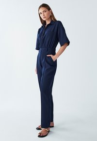 Marineblauwe jumpsuit met korte mouwen, ritssluiting aan de voorkant, twee borstzakken en een gecinchde taille. Gladde stof en taps toelopende benen. Draagt met platte sandalen.