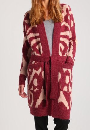 Femme portant un long cardigan en maille à motifs bordeaux et beige avec une ceinture et des poches, par-dessus une chemise grise et un pantalon noir.