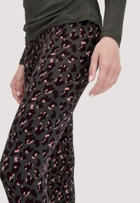 Vrouw in donkere leggings met een roze en zwarte panterprint, gecombineerd met een langwerpige donkergrijze top, hand ontspannen aan de zijkant.