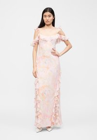 Γυναίκα με μακριά μαύρα μαλλιά που φοράει παστέλ μακριά φόρεμα με λουλούδια, με αποκαλυπτικά ώμους και βολάν στο τελείωμα, στέκεται με το ένα χέρι στη μέση, φαίνονται λευκά τακούνια.