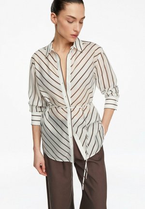 Frau trägt ein transparentes weißes Hemd mit braunen diagonalen Streifen, in der Taille gerafft, kombiniert mit lockeren braunen Hosen.