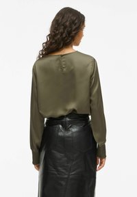 Vrouw met lang, krullend haar, gekleed in een olijfgroene blouse met lange mouwen en een zwarte leren rok, gezien van achteren.