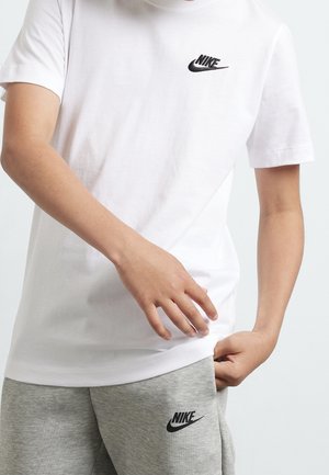 Personne portant un t-shirt blanc Nike et un short gris Nike, une main ajustant le t-shirt et l'autre dans la poche.