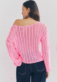 Maglione rosa lavorato a maglia con un design a trama aperta, scollatura ampia e maniche lunghe, abbinato a pantaloni in denim scuri. Aspetto textured e leggero.