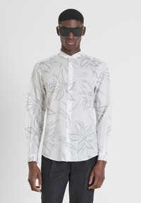 Camisa blanca de manga larga con patrón de hojas grises, cuello abotonado y botones en la parte delantera. Combinada con gafas de sol negras y pantalones oscuros.