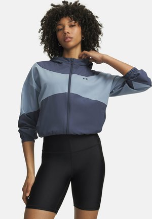 Frau mit lockigem Haar, die eine blaue Colorblock-Zipjacke und schwarze Radhosen trägt und vor einem einfarbigen Hintergrund steht.