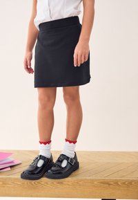 Jupe en coton noir à la texture lisse, portée avec des chaussettes blanches ornées d'accents rouges en forme de cœur et des chaussures à boucles en cuir verni noir.