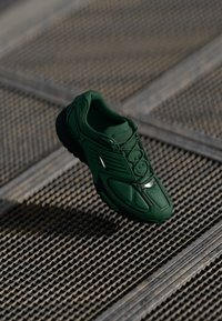 Sneaker Nike vert foncé avec semelle noire et panneaux détaillés, reposant sur une surface en métal texturé avec des jeux de lumière et d'ombre diagonaux.