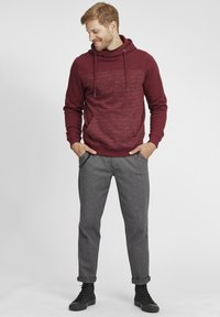 Felpa bordeaux con una sezione superiore testurizzata, cordini e tasche; abbinata a pantaloni grigi testurizzati e sneakers nere.