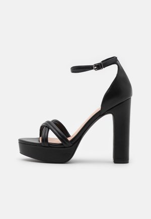 Sandale noire à talons hauts avec plateforme, présentant plusieurs brides entrecroisées et une sangle de cheville maintenue par une boucle en métal.