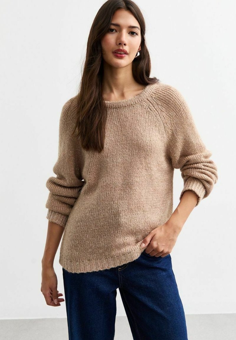 Beige stickad tröja med rund halsringning, sänkt axlar och tjocka muddar, parat med blå jeans, fotograferad mot en enkel bakgrund.