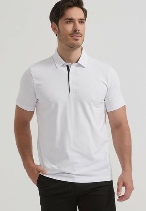 Homme portant un polo blanc à manches courtes avec col et pantalon noir, debout avec une main dans la poche sur un fond uni.
