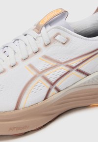 Weißer Sportschuh mit strukturiertem Obermaterial, braunen und gelben Akzenten sowie einer gepolsterten Sohle. Verfügt über ein Logotag und geflochtene Schnürsenkel.